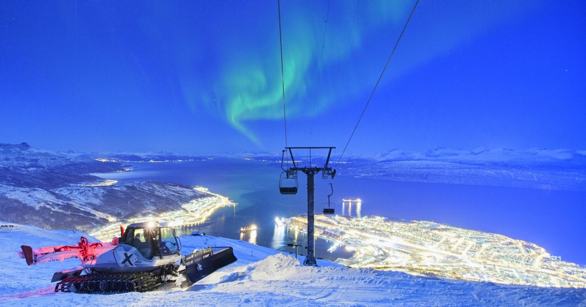Nordlys over skiheisen i narvikfjellet og preppemaskin. Lyset fra byen ses i bakgrunnen. Foto