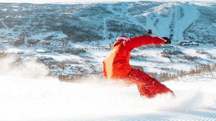 En person i røde klær som suser nedover en preparert skibakke, med snøsprøyt rundt seg. Foto.