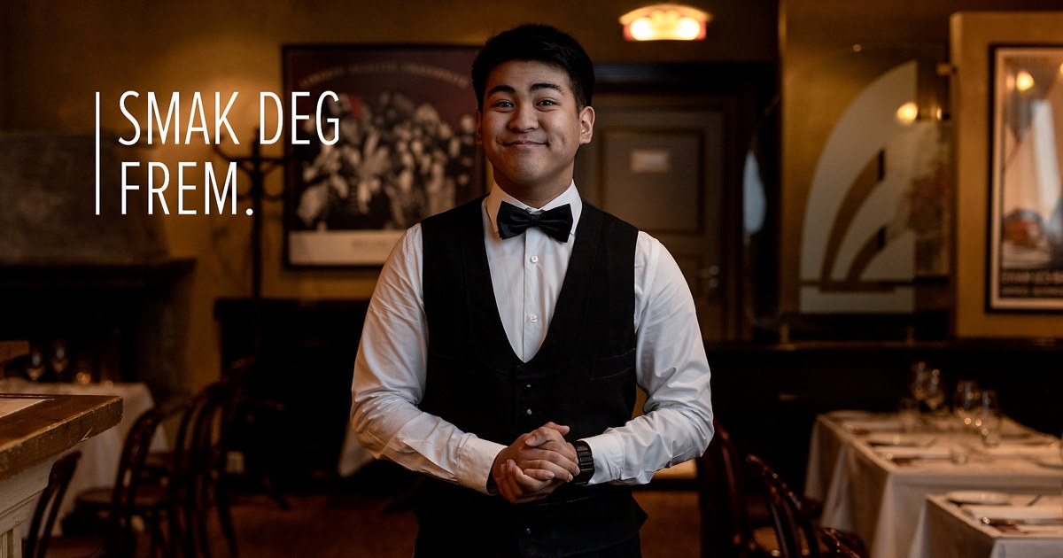 Ung, smilende mann i servitøruniform som står i en restaurant. Foto. Over bildet står teksten "Smak deg frem".