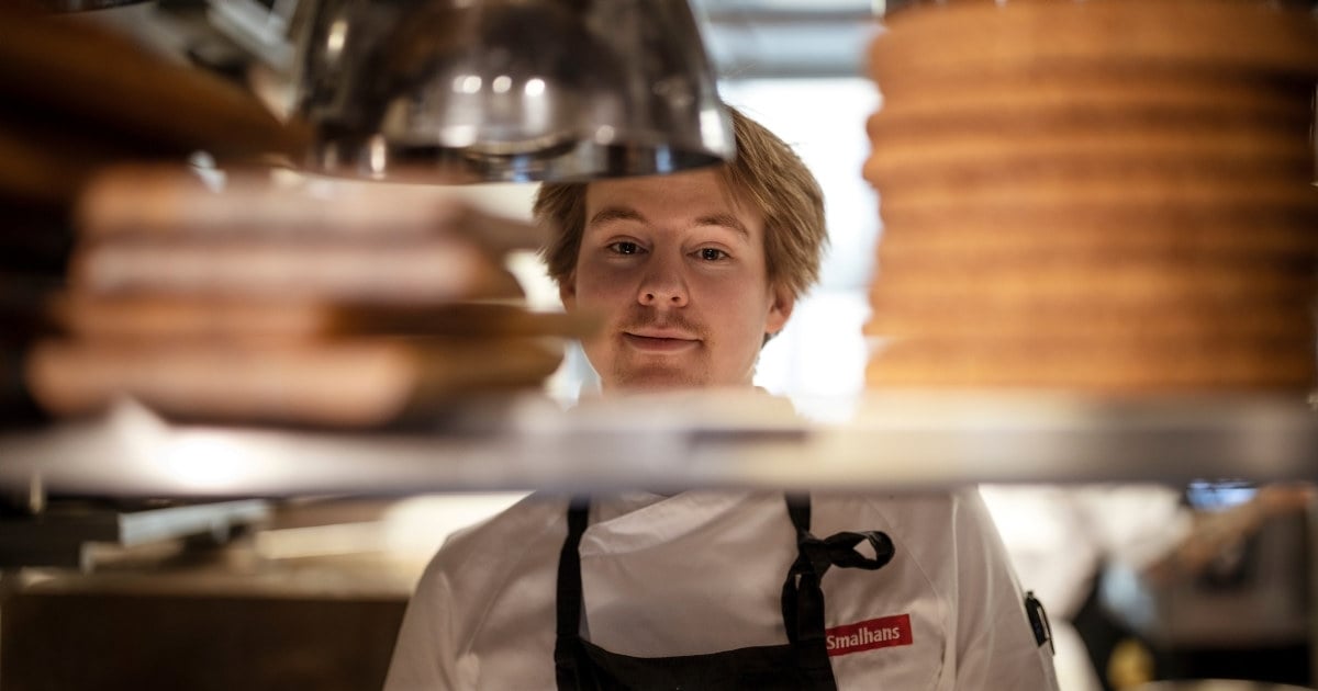 Ung kokk som titter mellom serveringshyller på et restaurantkjøkken. Foto