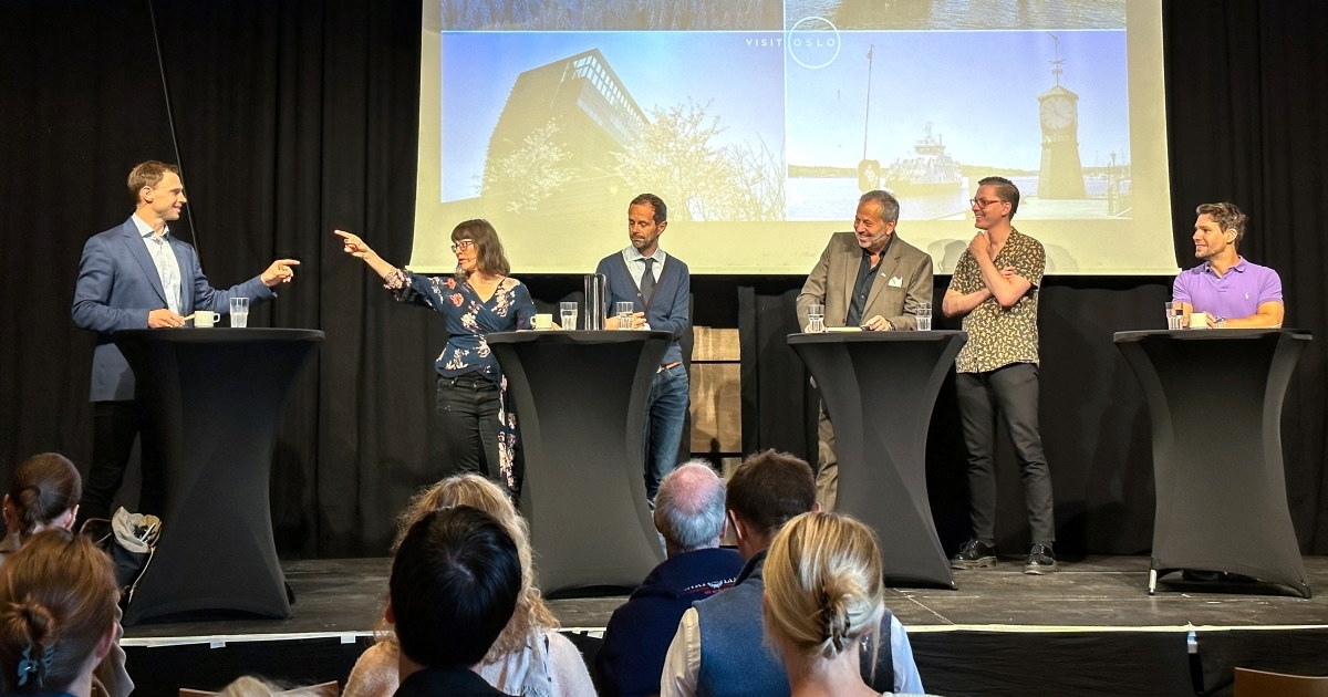 Seks smilende mennesker som står på en rekke på en scene med en presentasjon i bakgrunnen. Mannen og kvinnen som står ytterst til venstre i rekken peker på hverandre.