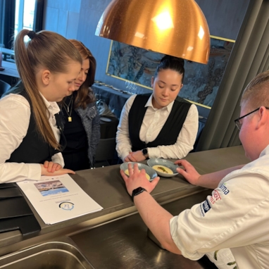 Tre servitører og en kokk som står ved anretningen, der det står to retter. Foto