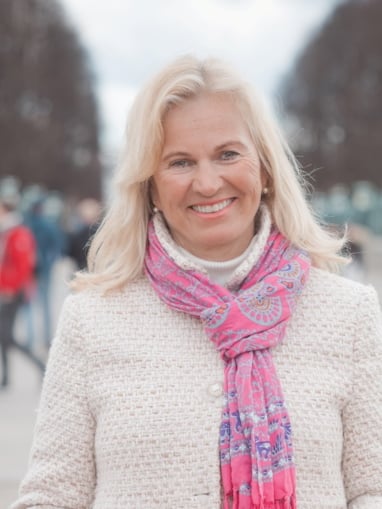 En kvinne i lys jakke og rosa skjerf som smiler mot kamera. Foto.