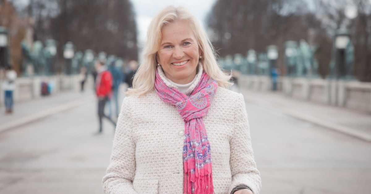En kvinne i lys jakke og rosa skjerf som smiler mot kamera. Foto.