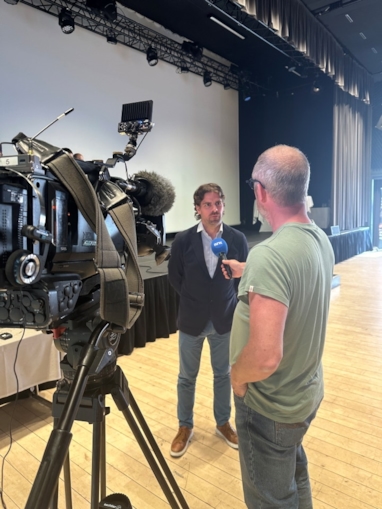 Mann kledd i dresjakke som intervjues av en mann i grønn t-skjorte med et TV-kamera i forgrunnen.