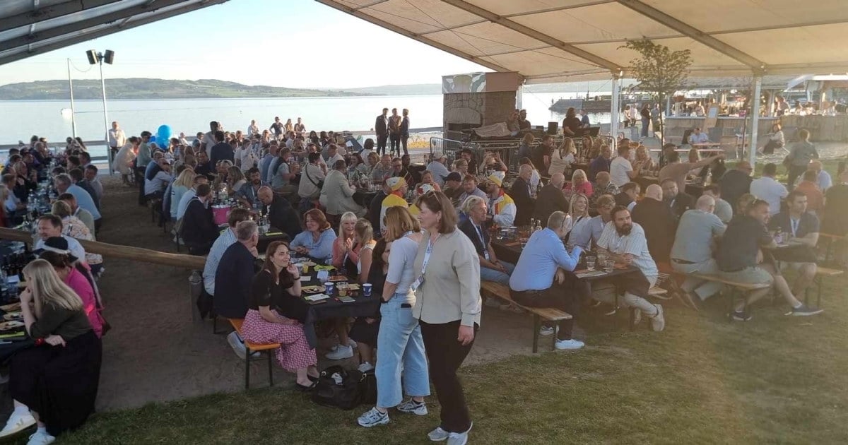 Mange mennesker som sitter ved langbord under et stort telttak. Foto